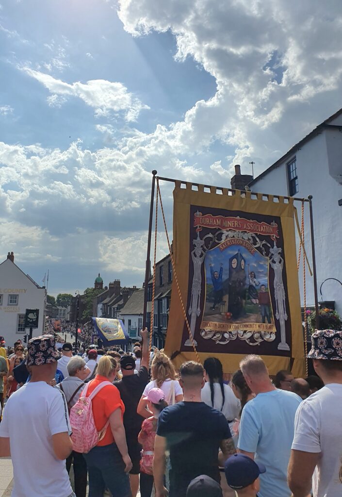 People marching down the street and carrying the Durham Miners' Association banner at the 2023 Durham Miners' Gala parade.