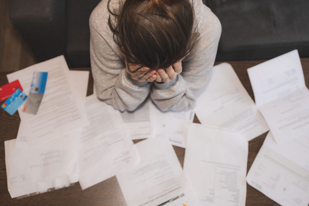 This photo shows an overhead view of a women who is sitting at a desk - she is looking down with her hands covering her face and seems stressed. The desk is covered with sheets of paper that look like bills and there are 4 credit cards on top of the papers near the woman's left elbow.